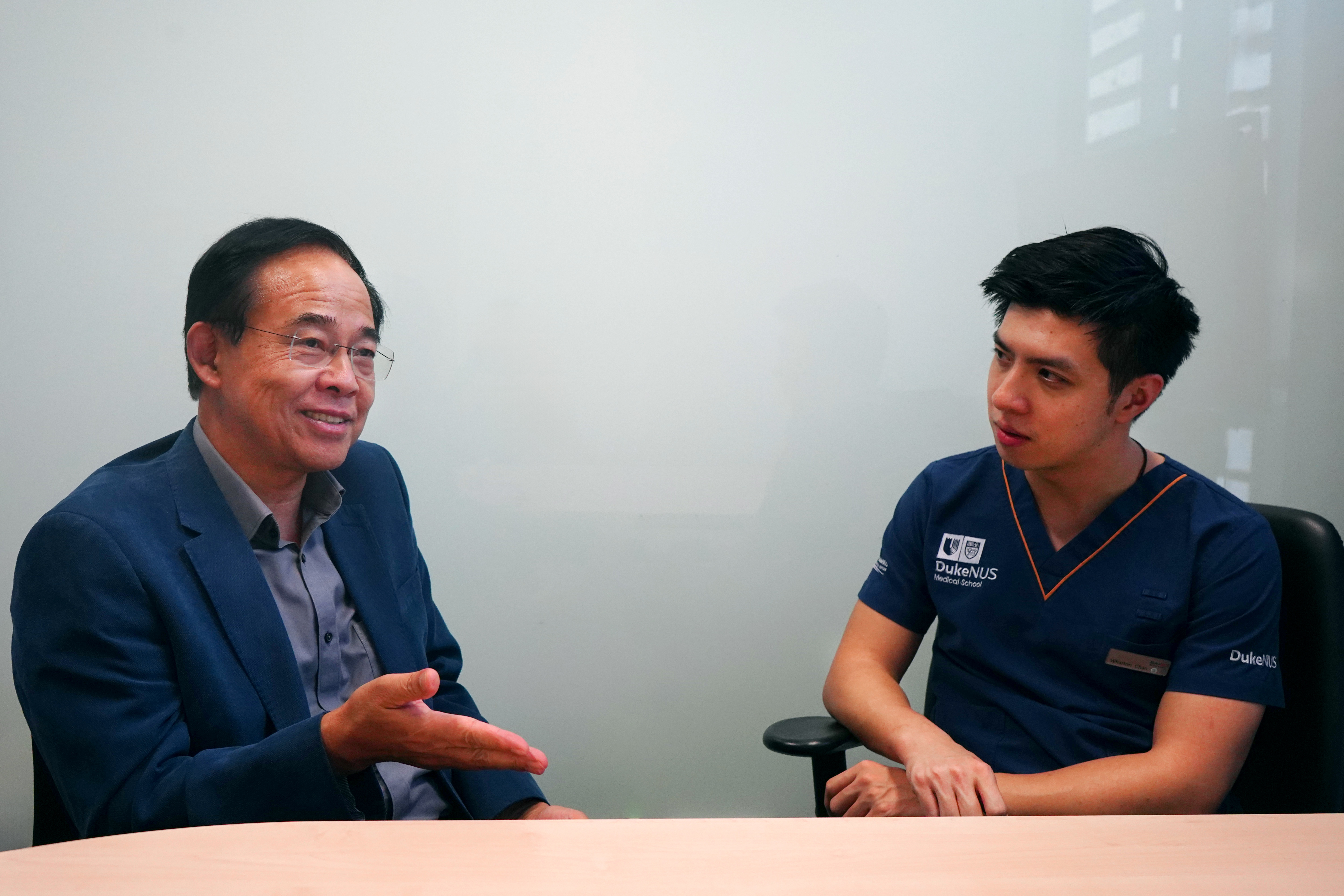 A mid-body shot of Professor Wang Linfa and Dr Wharton Chan, left to right, sitting at a table in dark blue medical scrubs and having a conversation.