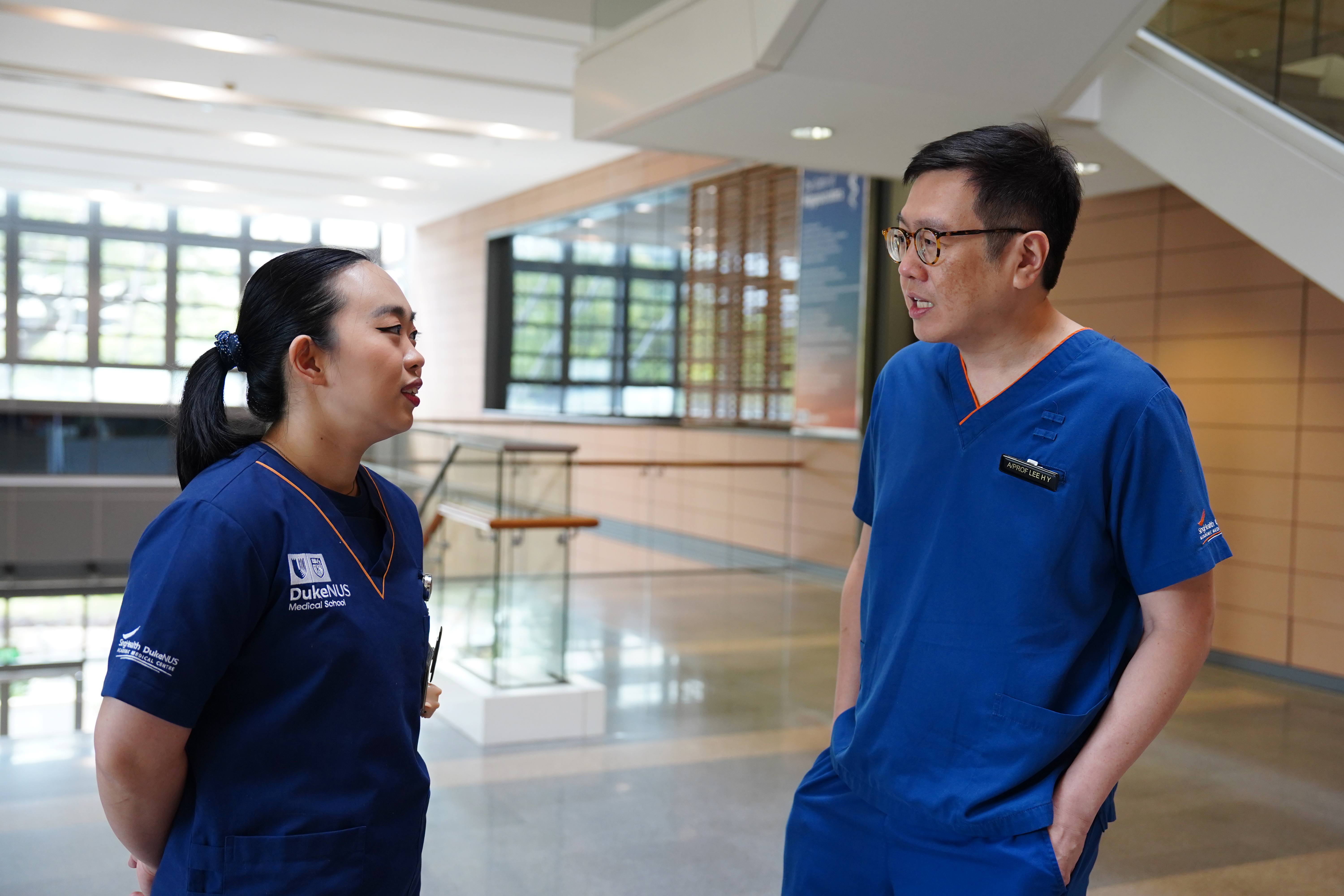A medium shot of Ms Esther Seow, on the left, and Professor Lee Haur Yueh, on the right, facing each other and laughing while having a conversation, in medical scrubs.