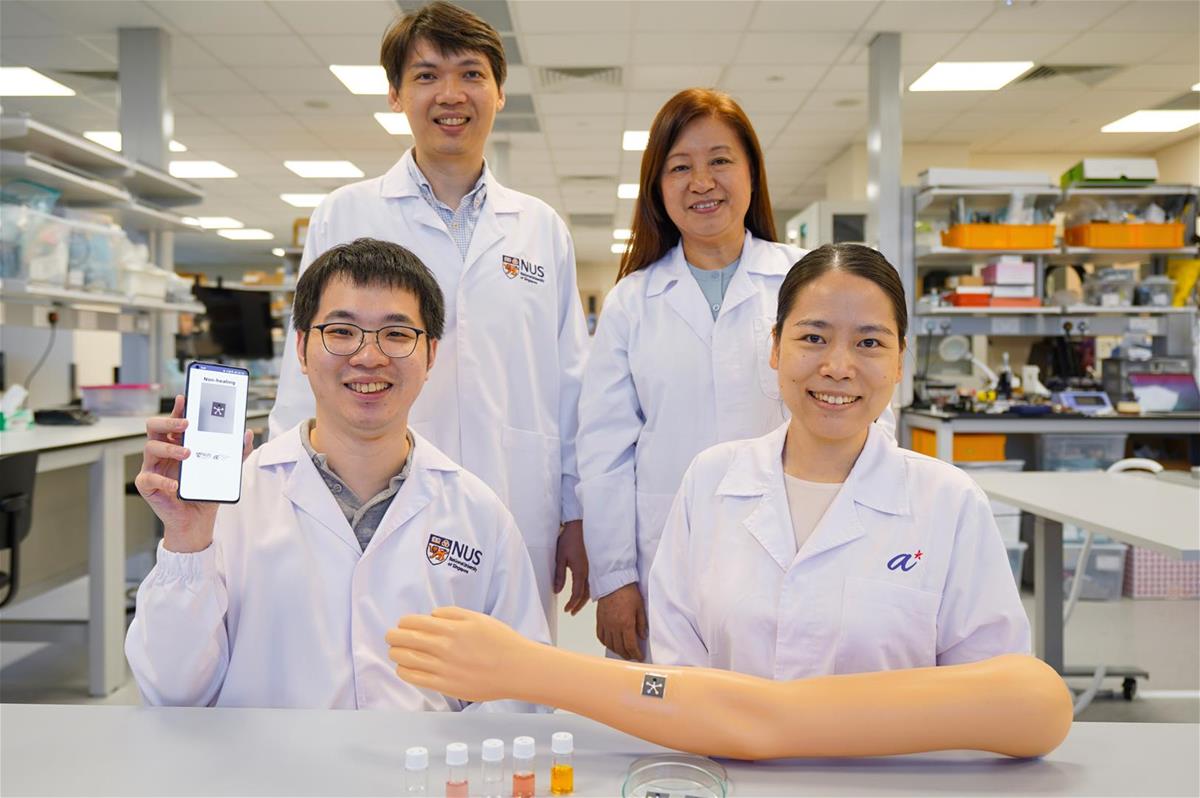 A team of four scientists, one holding up a mobile phone, gathered behind a false  arm with a PETAL sensor patch on it