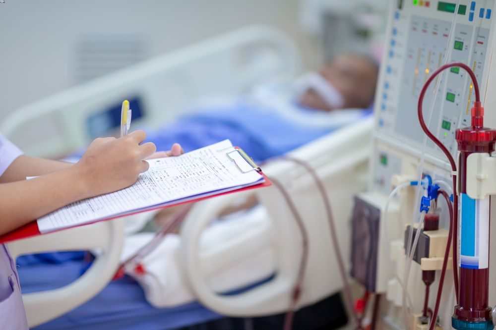 A stock photo of a dialysis nurse checking dialysis machine with a patient in the background